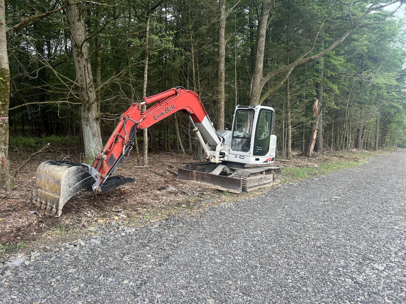 An excavator parked beside a gravel lane through the woods.